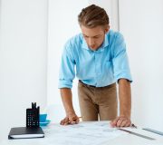 Picture of young handsome confident pensive businessman working standing at table drawing sketch. White modern office interior background.